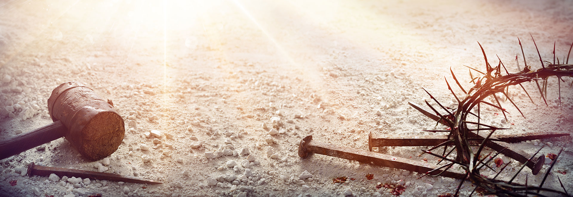 an old wooden mallet, rusted old nails, and a crown of thorns with drops of blood laying on rocky, sandy ground