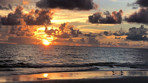 the sun setting into the ocean with numerous puffy white clouds